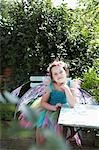 Portrait of young girl (5-6) in fairy costume sitting at garden table