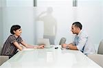 Man and woman talk over conference table, man on mobile phone visible through translucent office wall