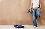 Woman with toolbelt and drill leaning against wall, low section