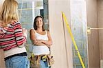 Woman with toolbelt leaning against wall, taling to friend