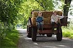 Two boys (5-6, 7-9) sitting on back of trailer on country lane