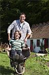 Father pushing son (7-9) in wheelbarrow outside cottage, portrait