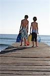 Two boys (6-11) walking on jetty carrying fishing nets and swim fins, back view