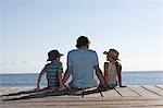 Father and two children (7-9) sitting on jetty, back view