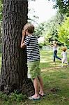 Boy (5-6) standing by tree with eyes covered, children running in background