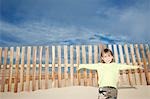 Garçon (5-6) adossé à une clôture en bois sur une dune de sable, portrait