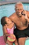 Senior homme avec GSM, assis près de la piscine avec une petite fille (5-6), portrait.