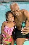 Senior man using mobile phone, photographing self with granddaughter (5-6) at swimming pool.