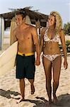 Surfer couple holding hands, carrying surfboard, walking on beach