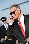 Mid-adult businessman talking on phone, airline pilot standing in background.