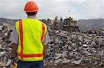 Worker watching digger moving waste at landfill site