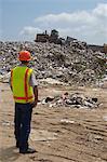 Worker watching digger moving waste at landfill site