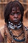 A Hamar woman in the village square of Dimeka. Married women wear two heavy steel necklaces. This woman wears an extra necklace with steel a steel phallic symbol which identifies her as a first wife. She wears her hair long in a braided fringe matted with animal fat and ochre.