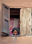 Une jeune fille peeps par la fenêtre à l'étage de sa maison près de Ambositra, Madagascar