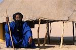 Crouching blue clad Tuareg elder outside his reed woven shelter