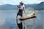 Myanmar. Burma. Lake Inle. An Intha fisherman with a traditional fish trap uses an unusual leg-rowing technique to propel his flat-bottomed boat across the lake while standing.