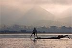 Myanmar,Burma,Lake Inle. An Intha fisherman with traditional fish trap uses an unusual leg-rowing technique to propel his flat-bottomed boat across the lake while standing.