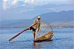 Myanmar, Burma, Lake Inle. Ein Intha-Fischer mit eine traditionelle Fischfalle verwendet eine ungewöhnliche Bein Rudern um seine flache Boot über den See stehen zu treiben.