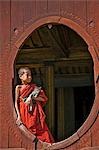 Myanmar. Burma. Nyaung-shwe. A young novice monk holding a cat at an oval window of the attractive wooden thein of the mid-19th century Shwe Yaunghwe monastery.