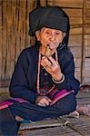 Myanmar,Burma,Wan doi. An old woman of the small Ann tribe in traditional attire sitting on the raised platform of her house as she smokes a pipe.