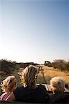 Namibia, Damaraland, Region Erongo. Ein junger Familie sitzen auf der offenen Plattform von einer Safari Allradantrieb ausgestattet, wie es entlang der sandigen Strecke schneiden durch Savanne und Bush fährt.