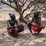 Two Turkana men in traditional attire relax in the heat of the day under a shady tree. Every man will have a wooden stool,which doubles up as a pillow at night to protect his clay hairdo. Men will never sit on the ground; only women and children are permitted to do so.