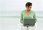 Woman sitting in chair using laptop, sea in background