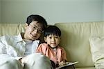 Father and son sitting on sofa with book, smiling at camera