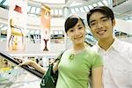 Couple taking escalator in shopping mall, smiling at camera