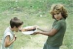 Boy holding up plate of grilled meat while second boy leans forward to take bite