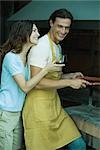 Couple standing by barbecue, woman holding glass of wine, both laughing