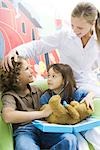 Boy and girl playing doctor with teddy bear while nurse watches