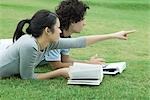 Young couple lying on grass with books, woman pointing, both looking out of frame