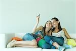 Young woman photographing self and friend with cell phone, sitting on couch