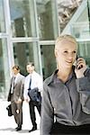 Young businesswoman using cell phone outdoors, looking away, businessmen walking in background