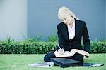 Young businesswoman sitting on the ground outdoors, reading book, holding pen