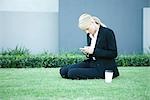 Young businesswoman sitting on the ground outdoors, looking at cell phone, hot beverage