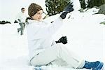 Adolescente assis sur le sol dans la neige, jetant des boules de neige, vue latérale