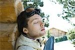 Young man leaning against log cabin, with snowboard, eyes closed