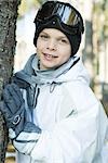 Boy wearing ski gear, leaning against tree trunk, smiling at camera, portrait