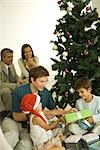 Father and two children sitting by Christmas tree, opening presents together, adults in background