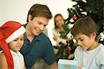 Vater und zwei Kinder sitzen am Weihnachtsbaum, Tochter tragen Nikolausmütze, Sohn Eröffnung anwesend