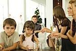 Two children holding Christmas ornaments, adults drinking champagne