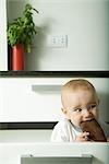 Baby in drawer, biting on wooden spoon, making funny face