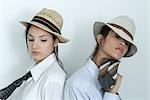 Two young female friends dressed in button down shirts, ties and hats, standing back to back, portrait