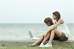Teen girl and little brother using laptop computer together by ocean, side view