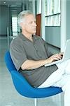 Mature man sitting in chair, using laptop computer, looking away