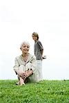 Senior woman sitting on grass, grandson standing behind her, holding gift, smiling, full length