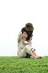 Senior woman sitting on grass, grandson leaning over her shoulder, handing her gift, full length