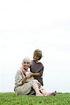 Senior woman sitting on grass, grandson handing her gift, full length